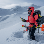 Backcountry skier holding an avalanche transceiver, probe and shovel in deep snow on a mountain slope