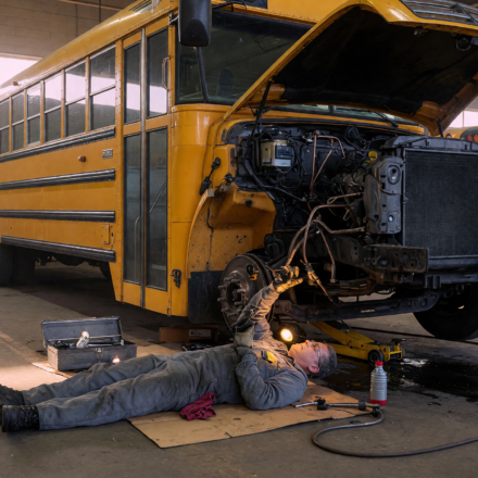 Yellow school bus in a maintenance depot with a mechanic inspecting the brake lines underneath