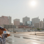 Hazy sun-baked city skyline with heat shimmer while a person shades their eyes and drinks water in extreme heat