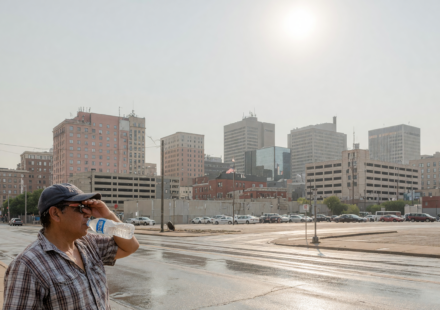 Hazy sun-baked city skyline with heat shimmer while a person shades their eyes and drinks water in extreme heat