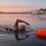 Open-water swimmer in a bright orange cap with a tow float in a calm lake at sunrise