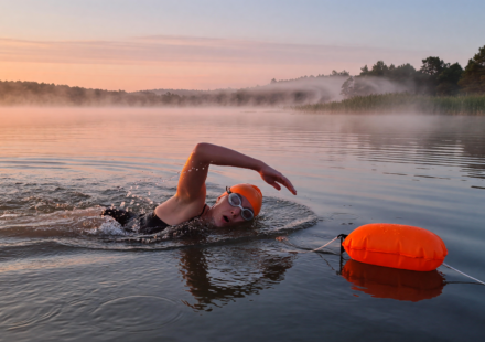 Open-water swimmer in a bright orange cap with a tow float in a calm lake at sunrise