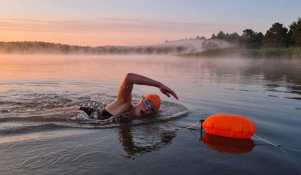 Open-water swimmer in a bright orange cap with a tow float in a calm lake at sunrise
