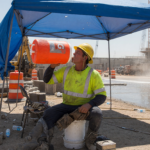Construction worker in a hi-vis shirt and hard hat resting in shade and drinking water from a cooler jug at a hot job site