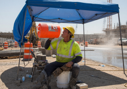 Construction worker in a hi-vis shirt and hard hat resting in shade and drinking water from a cooler jug at a hot job site