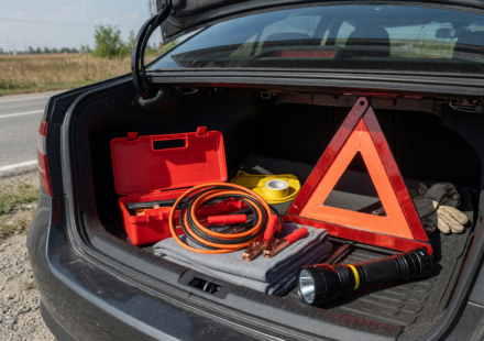 Open car trunk on a roadside showing an organized emergency kit with jumper cables, warning triangle and flashlight