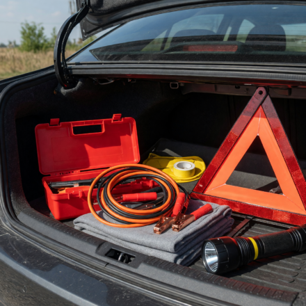 Open car trunk on a roadside showing an organized emergency kit with jumper cables, warning triangle and flashlight