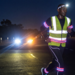 Runner at night on a dark road wearing a reflective vest and LED lights lit up by car headlights