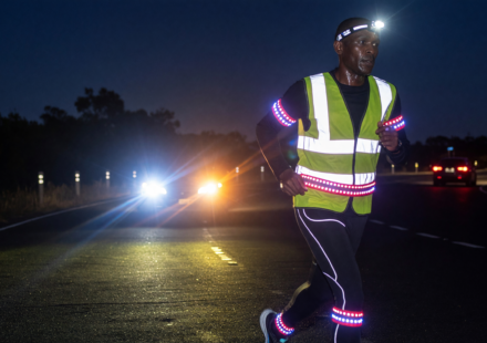 Runner at night on a dark road wearing a reflective vest and LED lights lit up by car headlights