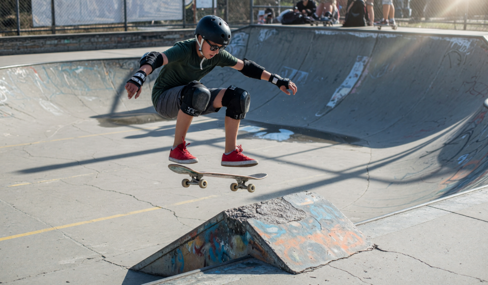 Skateboarder in helmet, wrist guards and knee pads mid-trick at a skatepark