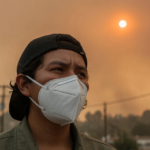 Person wearing a white N95 respirator outdoors under a hazy orange wildfire-smoke sky with a dim sun