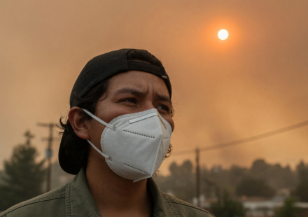 Person wearing a white N95 respirator outdoors under a hazy orange wildfire-smoke sky with a dim sun