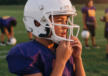 Youth football player on a practice field at golden hour adjusting the chin strap of a modern white football helmet