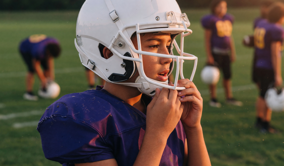 Youth football player on a practice field at golden hour adjusting the chin strap of a modern white football helmet