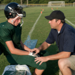 Youth athlete in a helmet on the sideline bench while a coach kneels to check on them