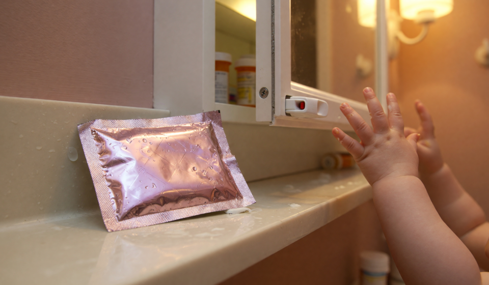 A foil numbing wipe packet on a bathroom counter beside a medicine cabinet with a child reaching in the background