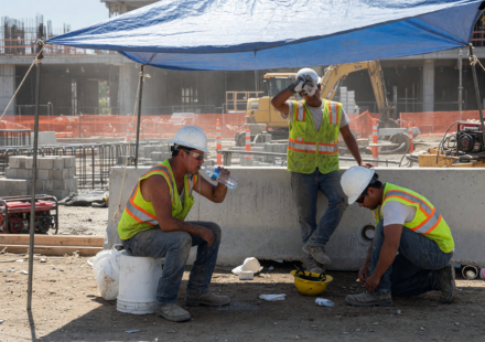 Construction workers in hard hats and hi-vis vests taking a shaded rest break and drinking water in bright summer heat