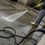 Person using an electric pressure washer on a wet driveway, with the power cord and plug near a wet outdoor outlet