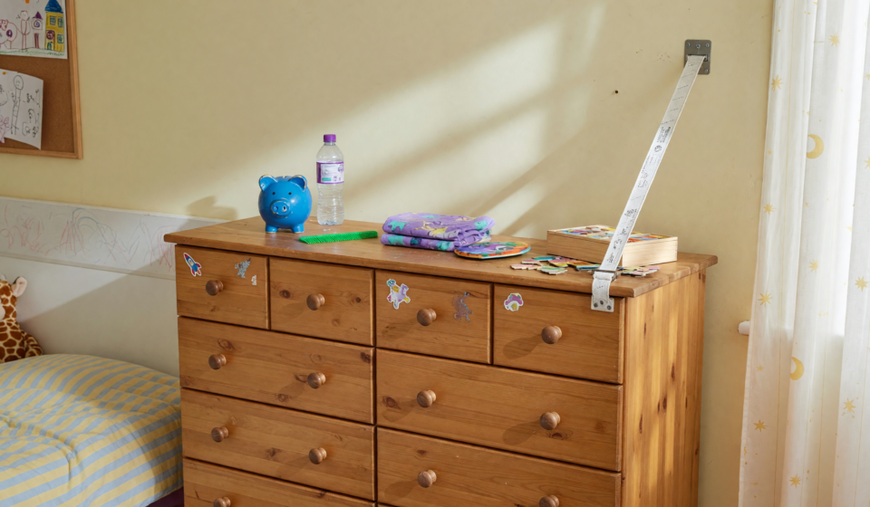 A wooden eight-drawer dresser secured to the wall with an anchor strap in a child's bedroom, illustrating tip-over prevention