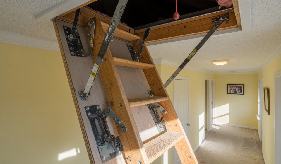 A pull-down attic stairway ladder unfolded into a home hallway, showing the hinge bolts and hardware at the center of the recall