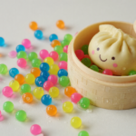 Colorful translucent water beads spilled beside a squishy dumpling-shaped novelty toy in a bamboo-steamer-style container