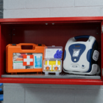 A workplace emergency cabinet holding a naloxone rescue kit and first-aid kit next to an AED, illustrating OSHA guidance on workplace opioid overdose readiness