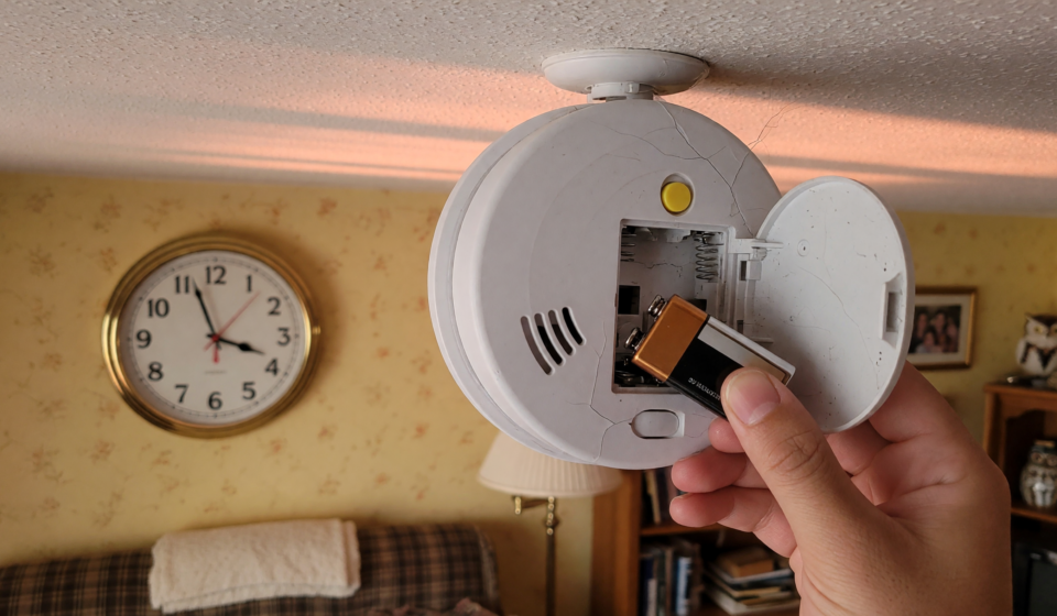 A hand inserting a fresh battery into a ceiling smoke detector as daylight saving time ends