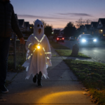A child in a Halloween costume holding a flashlight and wearing reflective tape walks with an adult on a suburban sidewalk at dusk